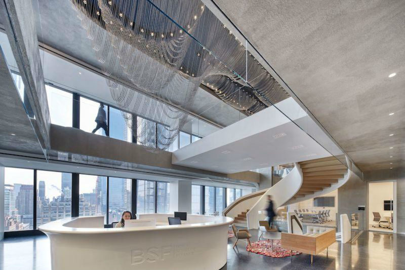 Boies Schiller Flexner

O design de escritórios de advocacia é geralmente pouco criativo - mas não nos novos espaços da Boies Schiller Flexner. Projetado pela Schiller Projects, o espaço de 9.290 metros quadrados, que abrange quatro andares, tem muito pouco em comum com a maioria dos escritórios do setor encontrados em Nova York.

Localizado na 55 Hudson Yards, o recinto parece ser o coworking mais sofisticado já construído - bem o oposto de um escritório de advocacia de alto nível. Os espaços de canto fechados foram substituídos por quartos semi-abertos com vistas panorâmicas do rio Hudson.

O espaço tem 22 salas para que os advogados trabalhem de maneira colaborativa, o que é uma parte importante da cultura corporativa da empresa. No entanto, chamadas de telefone podem ser feitas em uma das 24 salas para ligações privadas.

Para manter a produtividade no máximo, nenhum detalhe foi esquecido. O 19º andar tem até um café privativo de 370 metros quadrados e um café com serviço completo, o que dá aos funcionários menos um motivo para sair do prédio.

Carpetes e paredes brancas, que são típicos da maioria dos escritórios de advocacia, também não são encontrados por lá. Em vez disso, há piso de marmorite preto, assim como paredes de concreto curvo por toda parte.

A característica mais notável é “The Ribbons”, uma instalação de arte feita de contas metálicas que conecta as áreas comuns ao longo dos três andares. É adjacente a uma escadaria curva que não passa despercebida.