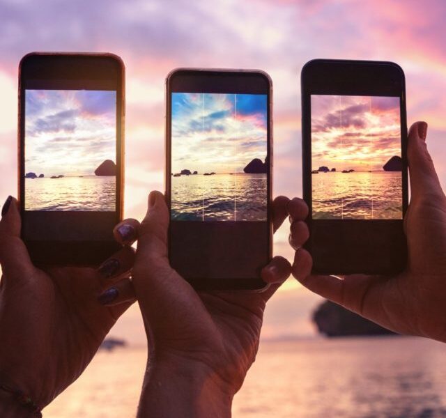 Três mãos segurando celulares enquanto fotografam o mar e céu