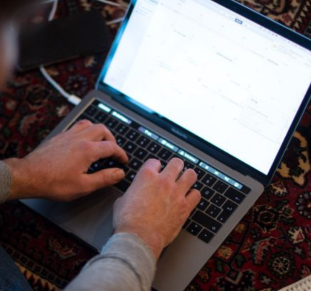 Pessoa digitando em notebook com carregador e celular ao lado