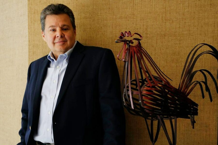 Joe Grendys, CEO da Koch Foods, de terno escuro, encostado em uma parede e ao lado de uma estátua de ferro de um galo