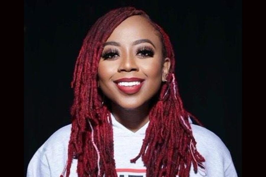 Aisha “Pinky” Cole, de cabelos vermelhos e trançados, maquiada, com largo sorriso. Veste um moleton branco