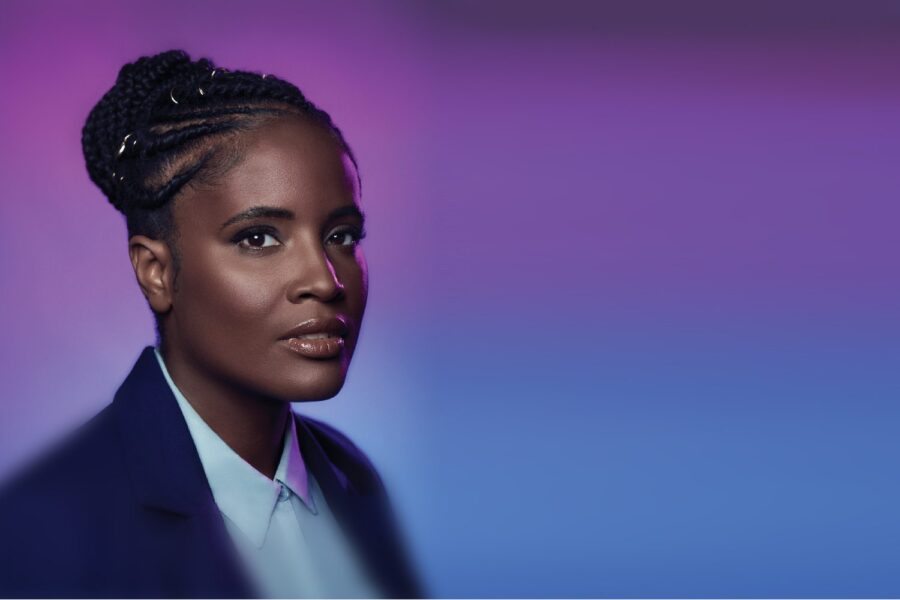 Djamila Ribeiro, mulher negra e de cabelo preso, à frente de fundo roxo