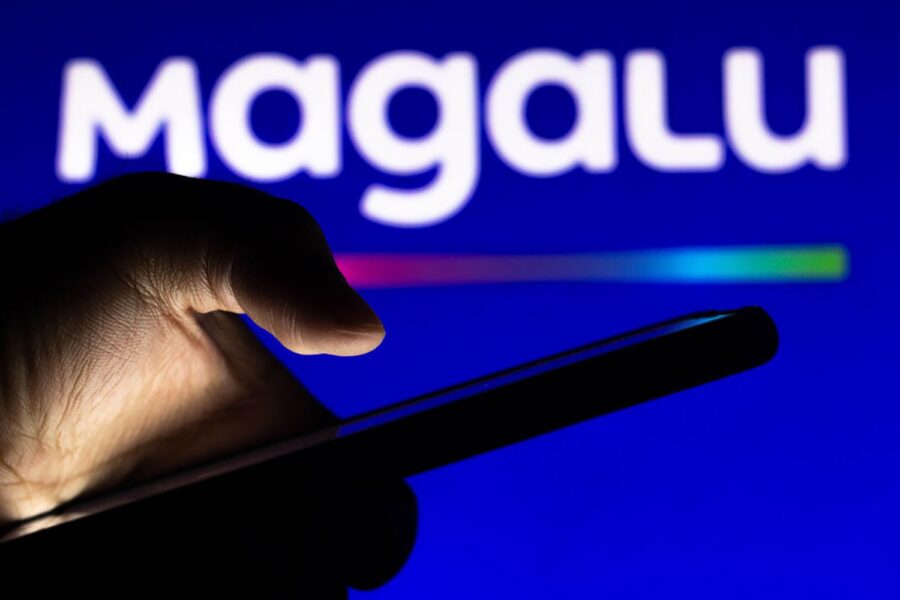 Pessoa com celular na mão na frente de tela com o logo "magalu" da Magazine Luiza