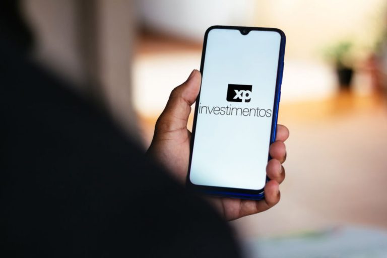 Pessoa com celular na mão e logotipo da XP Investimentos na tela