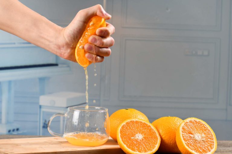 Mão espremendo meia laranja e ao lado do copo várias metades da fruta