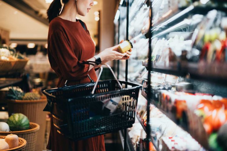 Mulher vestida de marrom, em frente a um refrigerador alimentos em um supermercado. Ela segura uma cesta, enquanto verifica um pote de alimento