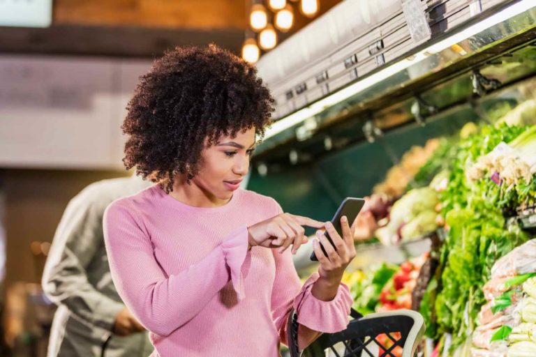 Mulher jovem e negra acessando um celular em frente a uma banca de vegetais de um supermercado