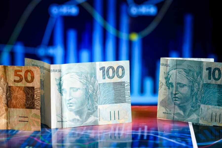 Duas notas de 100 reais e uma de cinco aparecem em frente a um gráfico