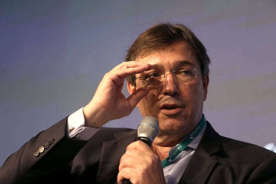 Wilson Ferreira Jr é um homem de pele branca, com cabelos castanhos. Ele usa óculos e segura um microfone durante um evento.