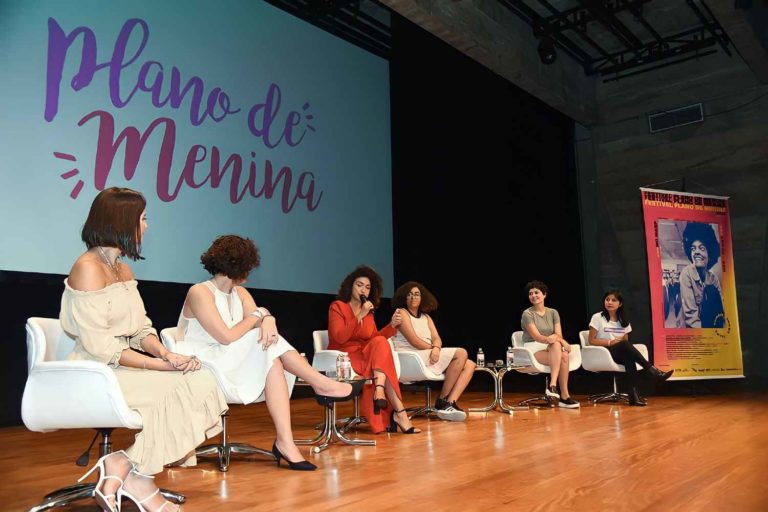 Seis mulheres sentadas conversando em um palco. No banner está o nome do festival Plano de Menina