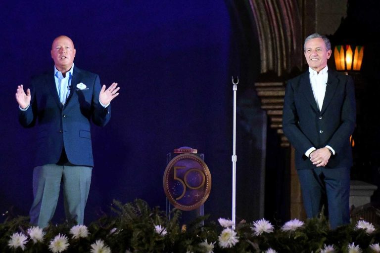 Dois homens de terno estão de pé em um palco, no qual há um microfone e um símbolo de 50 anos da Disney