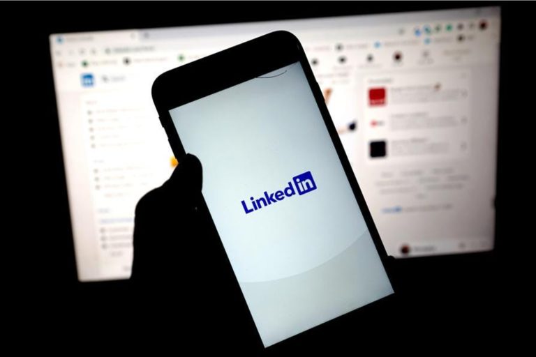 Pessoa segurando celular na mão com o logo do LinkedIn e, ao fundo, uma tela de computador com a página inicial da plataforma profissional