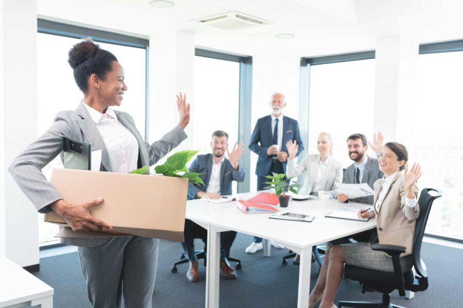 Mulher com uma caixa no braço se despedindo dos outros funcionários em empresa
