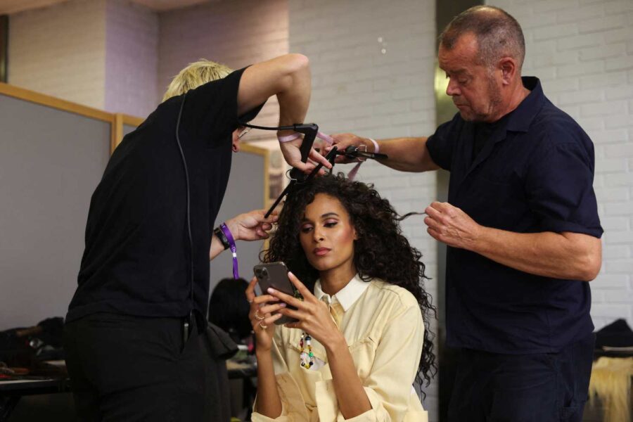 Mulher mexendo no celular tem seu cabelo arrumado por dois homens