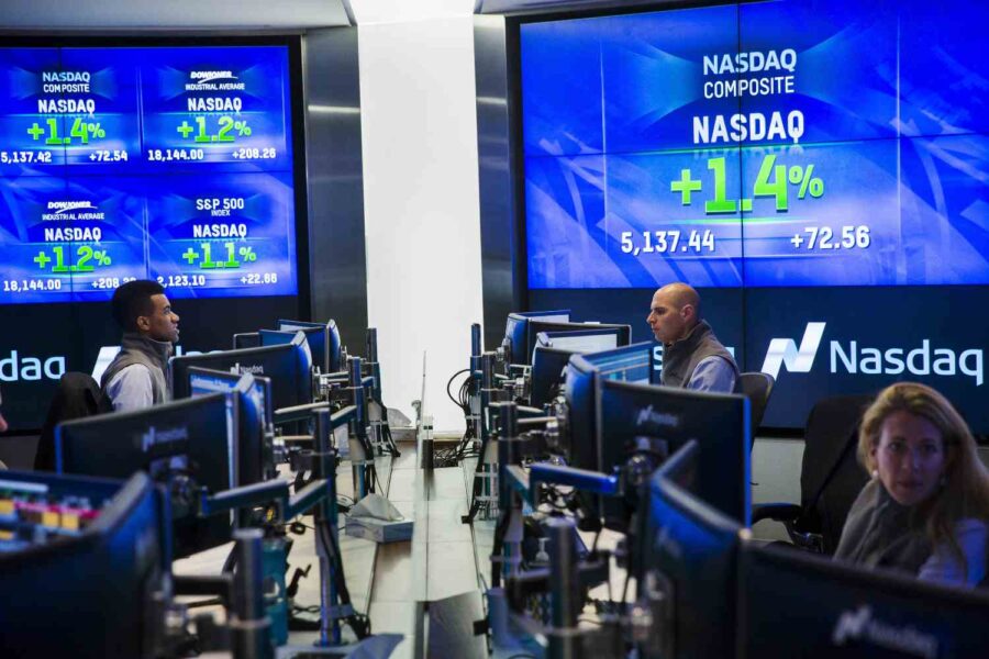Pessoas sentadas na frente de computadores e grades telas mostrando índices de ações ao fundo