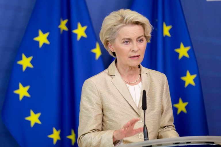 Mulher de terno falando em um microfone na frente de uma bandeira da União Europeia
