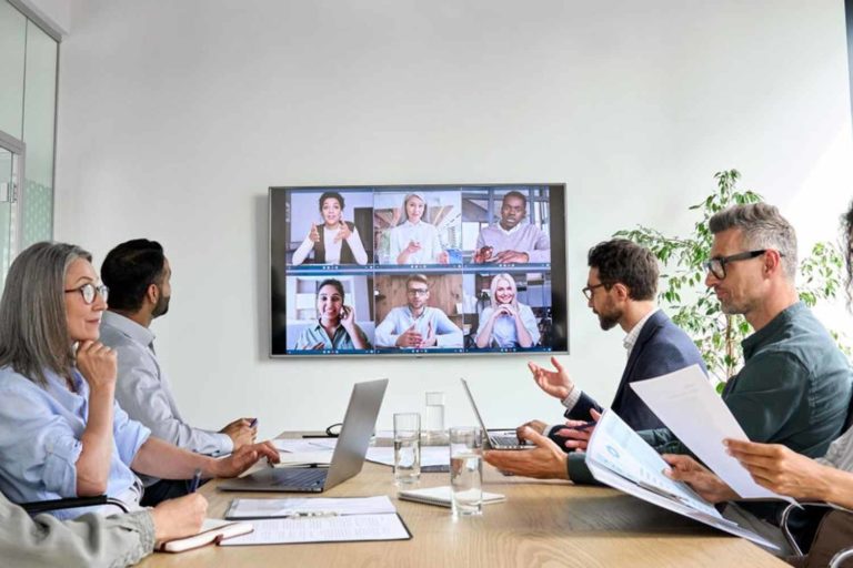 Pessoas sentadas em uma mesa de reunião e televisão com pessoas virtualmente em reunião