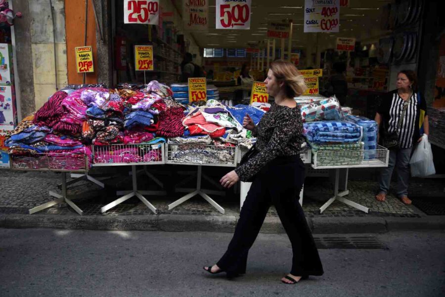 Mulher branca de cabelo loiro e roupas escuras caminha em frente à uma loja de roupas de comércio popular
