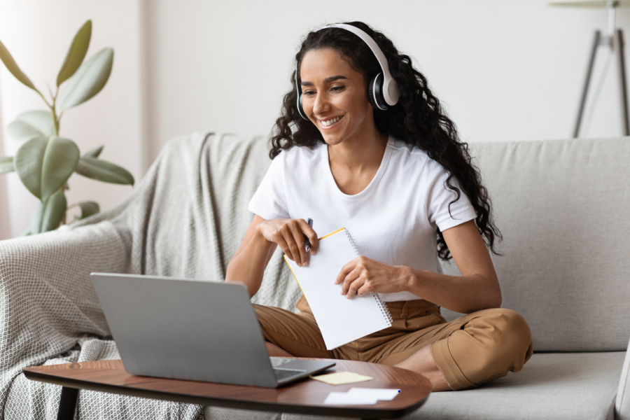 Mulher jovem sentada em um sofá, segurando um bloco de notas enquanto assiste uma aula remota em um notebook. Foto: Getty Images