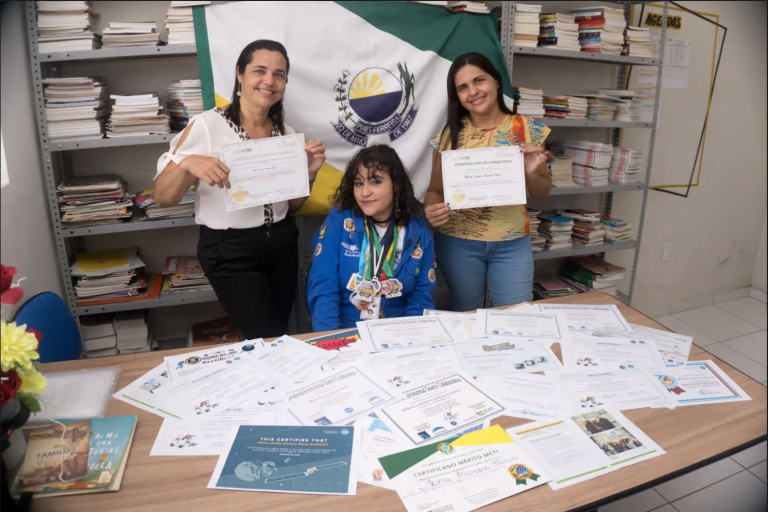 Larissa Paiva sentada ao lado de duas professoras em uma mesa repleta de certificados.