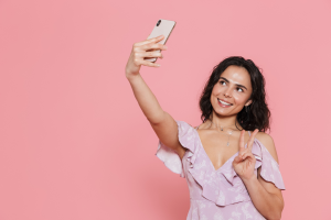 Mulher tirando uma "selfie" com um celular em um fundo rosa claro.