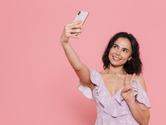 Mulher tirando uma "selfie" com um celular em um fundo rosa claro.