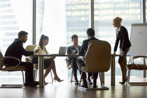 Foto de um ambiente corporativo, com pessoas sentadas em uma mesa ouvindo o que uma líder diz