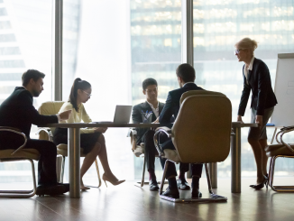 Foto de um ambiente corporativo, com pessoas sentadas em uma mesa ouvindo o que uma líder diz