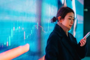 Mulher acompanhando gráficos do mercado financeiro em tela digital enquanto utiliza o celular.